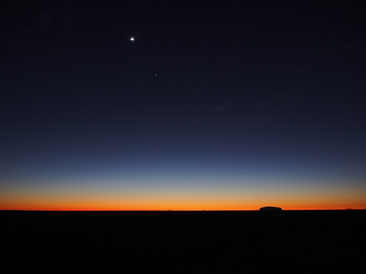 Der Mond am dunklen Himmel über Uluru, mit einem schmalen orangefarbenen Lichtstreifen am Horizont.