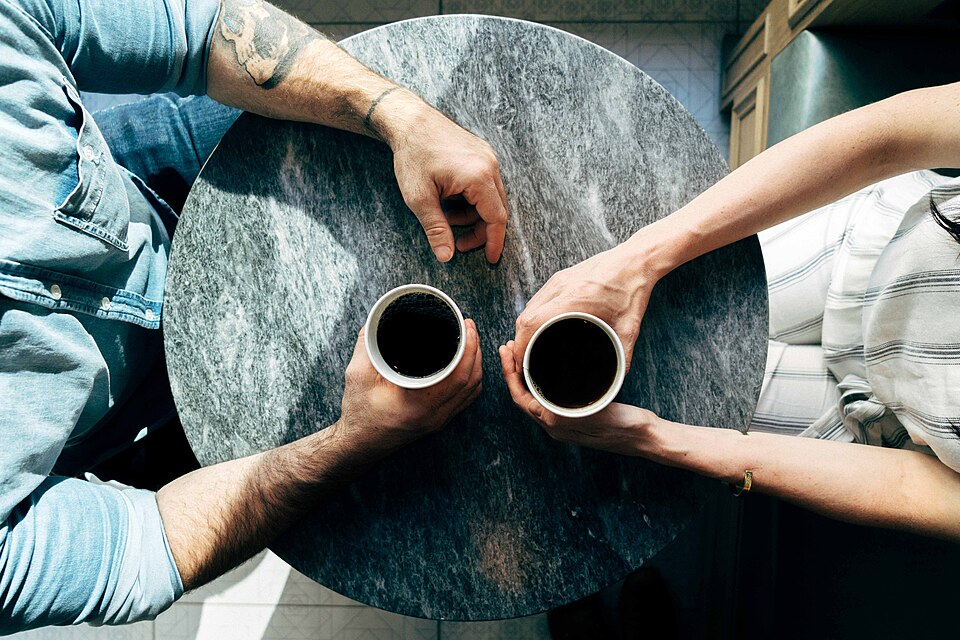 Zwei Menschen halten je eine Tasse schwarzen Kaffee an einem runden Tisch; nur Oberkörper und Hände sind zu sehen.